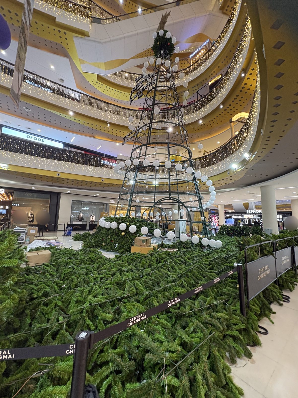 モールの吹き抜けで組み立て中のクリスマスツリー。骨組みに白い玉がつき、床には緑の枝がしきつめられている
