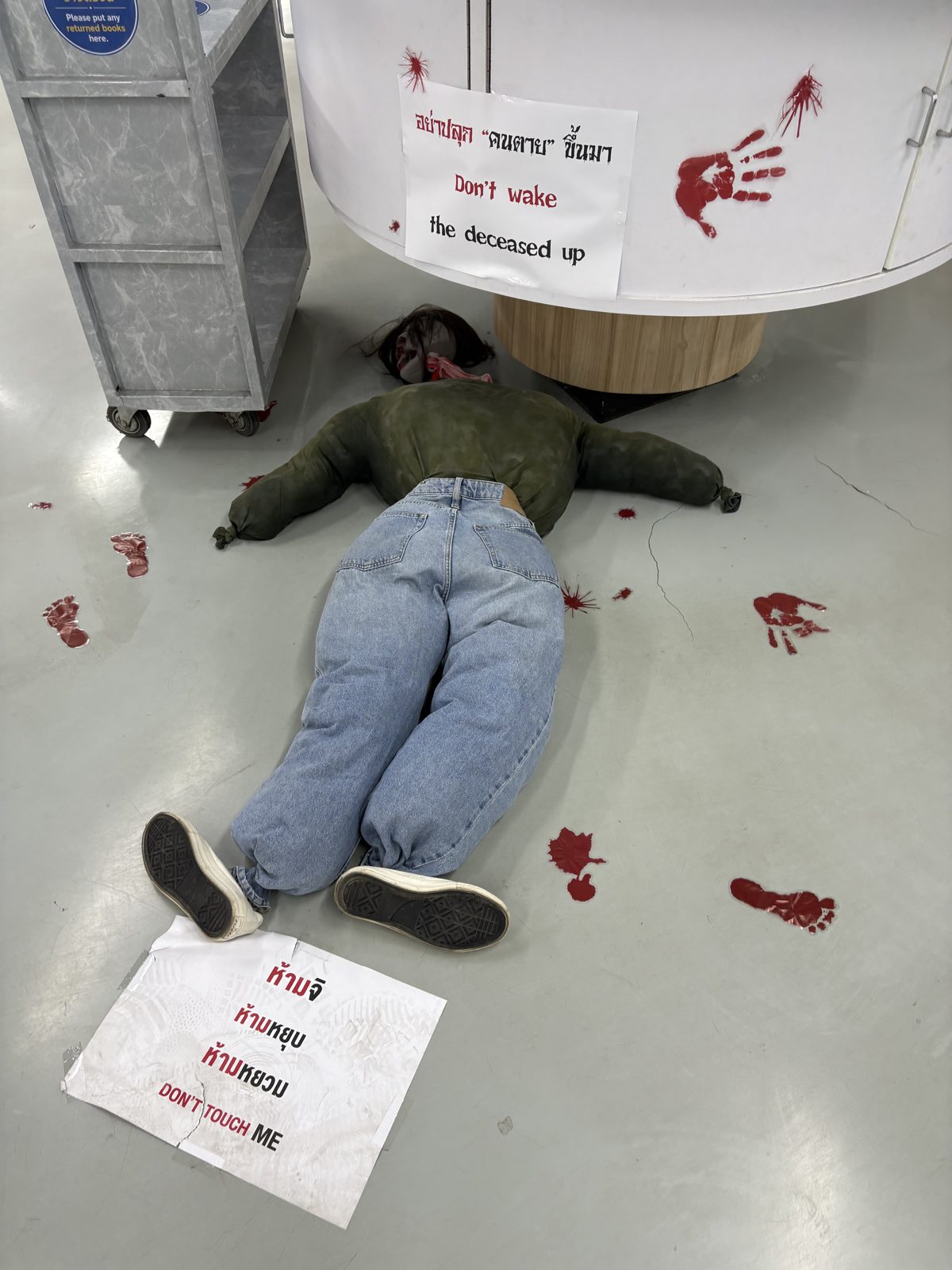 図書館の床にうつぶせに倒れた人形。まわりには赤い手形と足跡、「Don't wake the deceased up」「DON'T TOUCH ME」の張り紙